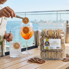 Person garnishing a cocktail with dried lemon slices, next to a package of dried lemon sticks and a glass of lemonade.